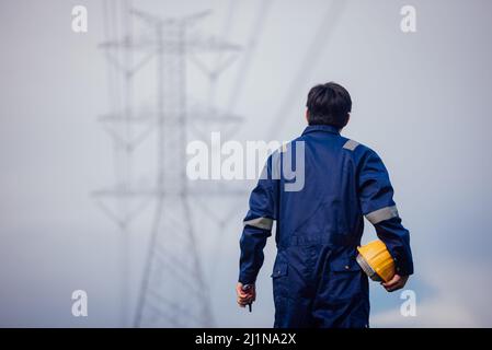 Un ingénieur d'alimentation mâle dans un casque jaune inspecte la ligne d'alimentation sous une ligne électrique haute tension. Banque D'Images