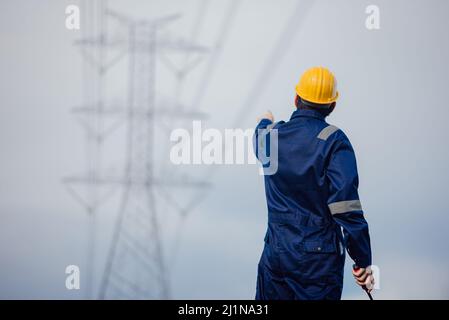 Un ingénieur d'alimentation mâle dans un casque jaune inspecte la ligne d'alimentation sous une ligne électrique haute tension. Banque D'Images