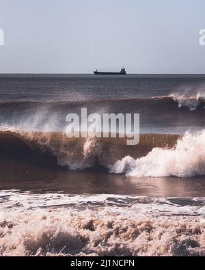 Les vagues de l'océan Atlantique se brisent près de la côte venteuse du Portugal et d'un petit pétrolier côtier ou navire-citerne loin à l'horizon. Composition verticale Banque D'Images