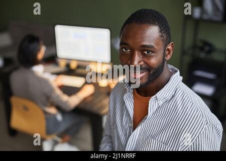 Portrait d'homme d'affaires noir moderne souriant à l'appareil photo tout en se tenant dans un espace de bureau avec des personnes en arrière-plan Banque D'Images