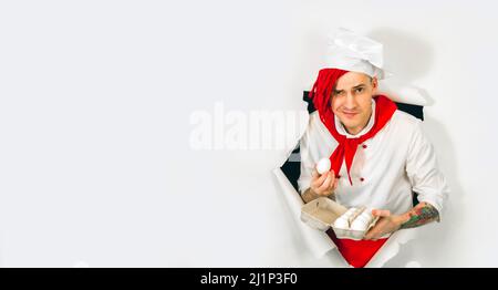 Un homme beau avec des dreadlocks rouges le tient dans ses œufs de poulet de main. Jeune cuisinier confiant en chemise blanche et cravate rouge tenant les oeufs de poulet, peeking th Banque D'Images