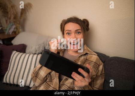 Happy girl étudiant joue un jeu vidéo sur une console de jeux portable à la maison, dans la chambre. Jeux en ligne avec des amis, jeux mobiles, prix, gagner, fun. À la sortie Banque D'Images