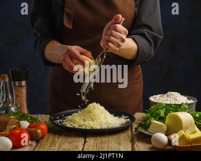 Un chef professionnel dans un uniforme sombre râpe du fromage sur une râpe à main sur une table en bois sur un fond bleu foncé. Beaucoup d'ingrédients pour pizza, tarte, Banque D'Images