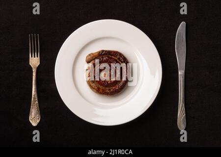 Saucisse en spirale sur une assiette blanche avec une fourchette et un couteau sur une nappe noire Banque D'Images