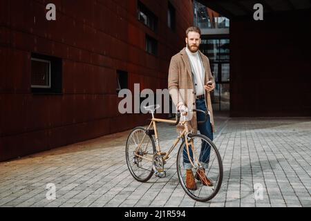 Jeune homme avec vélo tenant un smartphone devant le bâtiment regardant la caméra. Banque D'Images
