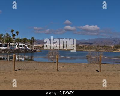 Vue sur le petit lagon de la Charca à Meloneras, une partie de Maspalomas, au sud de la Grande Canarie, en Espagne avec clôture en face et les touristes marchant sur la promenade. Banque D'Images