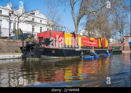 Deux jeunes femmes en paddle-board passent devant la Barge du théâtre de marionnettes sur le canal de la Grande Union à Little Venice. Londres, Angleterre, Royaume-Uni. Banque D'Images