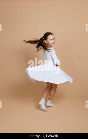 Portrait de jeune belle fille souriante mignonne avec de longs cheveux foncés en robe blanche, chaussettes et chaussures de gymnastique tourbillonnant en danse sur fond marron. Banque D'Images