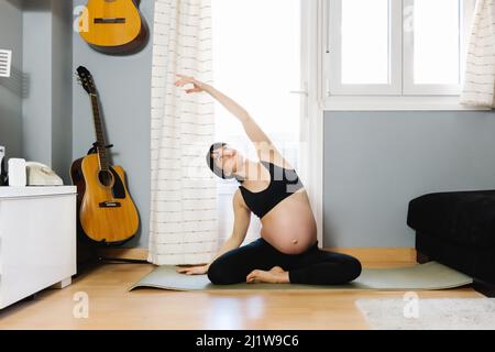 Femme enceinte de vêtements de sport assise sur un tapis et faisant une flexion latérale tout en pratiquant le yoga dans le salon Banque D'Images