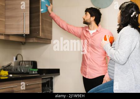 Vue latérale d'une jeune femme enceinte assise dans la cuisine et buvant du café chaud près d'un mari hispanique mettant une plaque en céramique dans le cabinet Banque D'Images