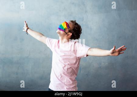 Anonyme transgenre couvrant le visage avec masque de protection coloré avec motif arc-en-ciel LGBT tout en se tenant debout avec les bras étalés sur fond gris Banque D'Images