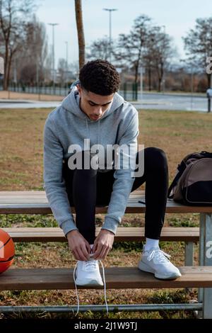 Corps complet de jeune homme hispanique dans des vêtements de sport nouant des lacets sur des baskets blanches tout en étant assis sur un banc en bois en ville Banque D'Images