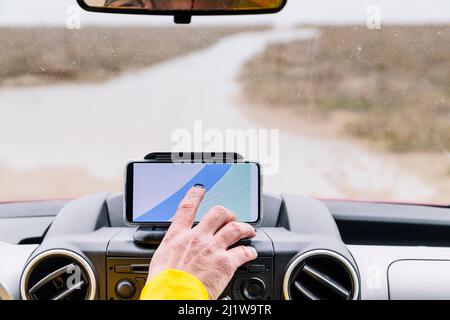 Main de crop chauffeur mâle anonyme tournant le navigateur sur le smartphone tout en étant assis dans une voiture garée sur la route dans la campagne Banque D'Images