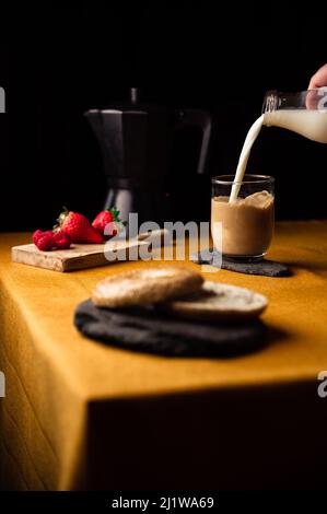 Croise la personne anonyme qui verse le lait de la carafe dans une tasse de verre avec du café infusé dans une marmite de moka pour le petit déjeuner avec des fraises et un bagel sur une assiette d'ardoise Banque D'Images