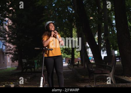 Une femme afro-américaine positive dans des vêtements décontractés attachant un casque de protection pour la conduite de trick électrique dans le parc parmi les arbres élevés Banque D'Images