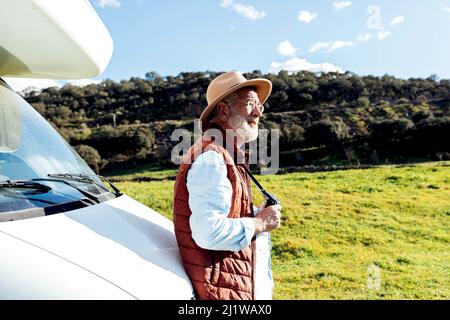 Vue latérale mâle senior touriste dans un chapeau tenant un appareil photo tout en se tenant près d'une voiture de camping garée sur un terrain herbacé tout en observant la nature le jour d'été Banque D'Images