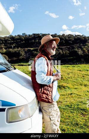 Vue latérale mâle senior touriste dans un chapeau tenant un appareil photo tout en se tenant près d'une voiture de camping garée sur un terrain herbacé tout en observant la nature le jour d'été Banque D'Images