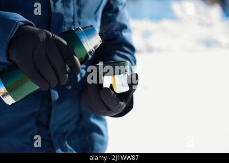 Crop-personne en manteau d'hiver bleu et gant chaud tenant une tasse en métal. Homme sans visage qui verse du thé chaud à partir de thermos pendant la journée froide d'hiver Banque D'Images