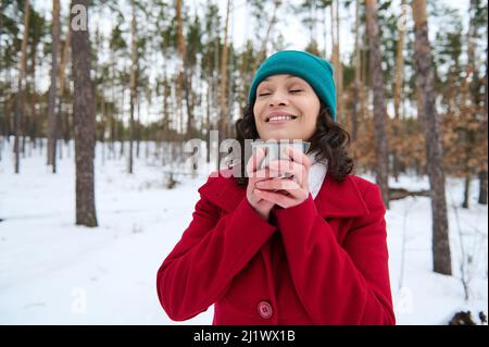 Une charmante femme caucasienne vêtue de vêtements chauds tient une tasse thermo avec une boisson chaude près de son visage, sourit avec un sourire éclatant en profitant d'un beau wi Banque D'Images