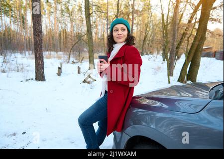 Charmante femme confiante dans des vêtements chauds se trouve sur le capot de sa voiture avec une tasse thermo avec une boisson chaude et apprécie sa pause café à l'extérieur sur un beau Banque D'Images