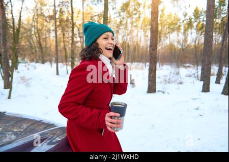 Une femme enjouée, assise sur le capot gris de la voiture et sourit en parlant sur un téléphone mobile, se détendant sur une forêt de pins enneigée avec une tasse thermo Banque D'Images