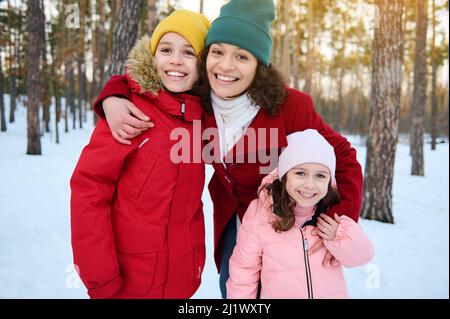 Bonne mère aimante - belle femme embrassant doucement ses adorables enfants, garçon et fille, souriant avec un sourire enjoué gai tout en marchant sur une couverture de neige Banque D'Images