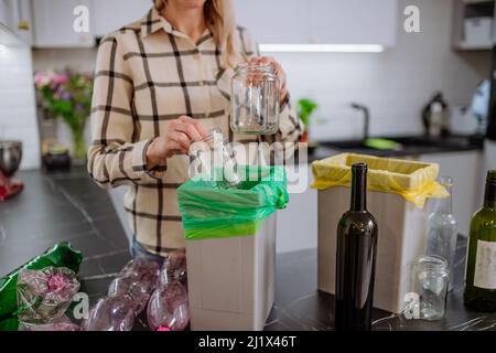 Section médiane de la femme jetant des bouteilles et des bocaux en verre vides dans le bac de recyclage de la cuisine. Banque D'Images