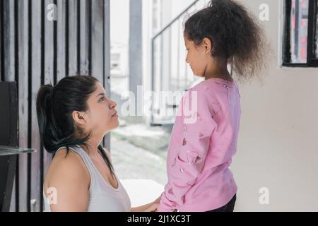 belle mère latina assise sur le sol tout en regardant sa fille à la peau brune dans les yeux, la fille est debout à côté de sa mère qui conseille Banque D'Images