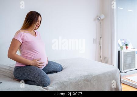 jeune femme blanche enceinte assise sur son lit avec les yeux fermés touchant son ventre, méditant seule se préparer à l'accouchement, concept de maternité, c Banque D'Images