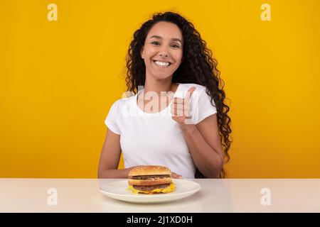Joyeuse Lady Holding Burger montrant Thumb Up Banque D'Images