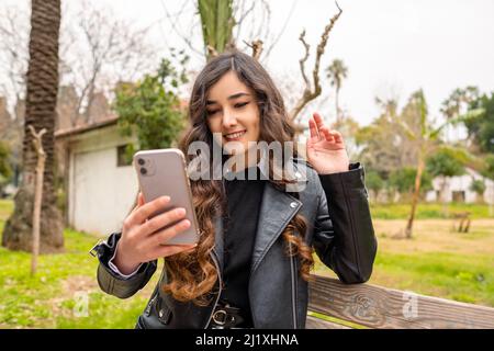 Jeune femme heureuse assise sur un banc de parc tenant un téléphone regardant l'écran de la main de la main vidéo d'appel distance ami en ligne mobile chat application. Smartphone Banque D'Images