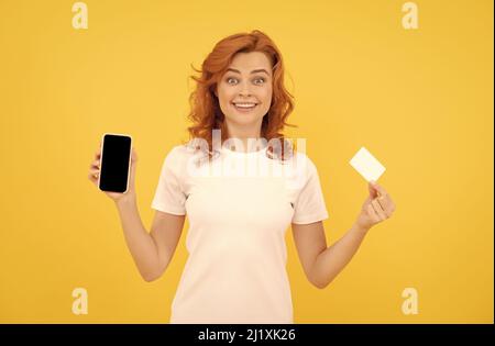 une jeune femme excitée a isolé un fond surjaune en utilisant un téléphone portable tenant une carte de crédit. Banque D'Images