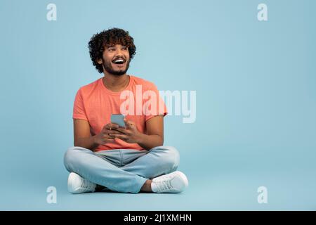 Un homme indien enthousiaste, assis sur un sol bleu, à l'aide d'un téléphone intelligent Banque D'Images