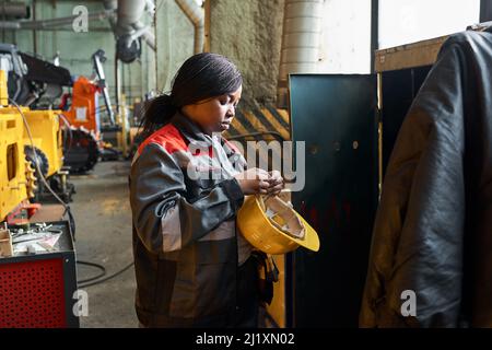Femme africaine en uniforme debout près de son placard et en train de retirer le casque de travail après son travail en usine Banque D'Images