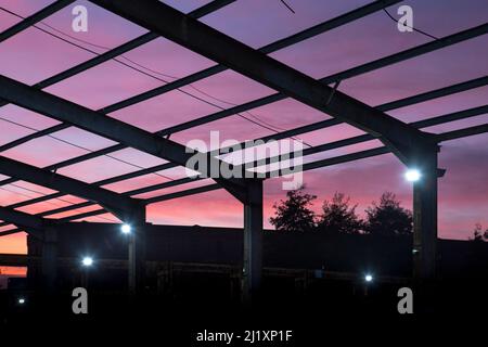 En regardant à travers une toile de poutres en acier, qui forit autrefois le toit d'un grand bâtiment avec le coucher de soleil coloré derrière et de petits projecteurs lumineux sur les montants verticaux. Banque D'Images