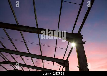 En regardant à travers une toile de poutres en acier, qui forit autrefois le toit d'un grand bâtiment avec le coucher de soleil coloré derrière et de petits projecteurs lumineux sur les montants verticaux. Banque D'Images