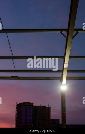 En regardant à travers une toile de poutres en acier, qui forit autrefois le toit d'un grand bâtiment avec le coucher de soleil coloré derrière et de petits projecteurs lumineux sur les montants verticaux. Banque D'Images