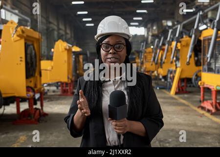 Portrait d'un journaliste africain en casque de travail tenant un microphone et parlant les nouvelles à la caméra tout en se tenant dans l'entrepôt Banque D'Images
