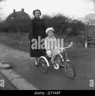 1950s, historique, à l'extérieur sur un trottoir de campagne, une mère avec sa fille assise sur son tricycle, Angleterre, Royaume-Uni. La dame tient une plomb attachée à l'arrière du tricycle, qui a été fait par TRIANG, un important fabricant britannique de jouets à base de métal pour enfants dans cette époque. Banque D'Images