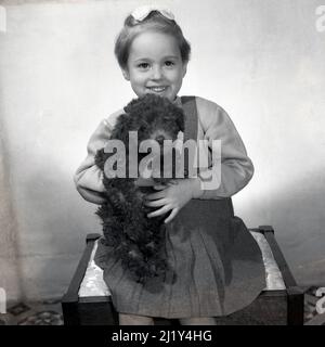 1950s, historique, une petite fille douce avec un noeud dans ses cheveux assis sur une petite table pour une photo tenant son chien d'animal de compagnie, une poddle, Angleterre, Royaume-Uni. Initialement élevé pour la chasse à la sauvagine, le Kennel Club a enregistré son premier poddle en 1874, avec un club de propriétaires fondé deux ans plus tard. Les chiens affectueux, les chiens amicaux, les poddles sont devenus en 1950s, l'une des races de chiens les plus populaires. Banque D'Images