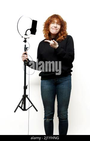 une femme aux cheveux rouges bouclés vêtue de jeans tient une ampoule avec une ampoule et un téléphone portable et la pointe avec un visage mécru sur une basa blanche Banque D'Images