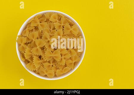 farfalle de pâtes dans un bol en céramique isolé sur fond jaune, pâtes brutes en forme d'arc, vue du dessus Banque D'Images