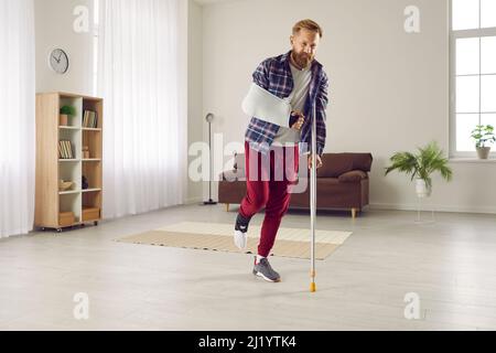 Homme malheureux avec jambe cassée et bras luttant avec la douleur et la marche avec la béquille à la maison Banque D'Images