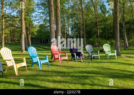 Une femme assise sur une chaise Adirondack avec des arbres et des bois en arrière-plan. Ombres. Banque D'Images