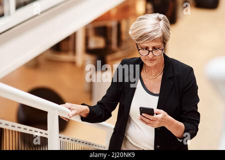 Faire semblant de passer des SMS en déplacement. Photo d'une femme d'affaires mature utilisant un téléphone portable tout en montant un escalier dans un bureau. Banque D'Images