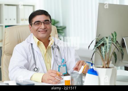 Portrait d'un médecin généraliste mûr souriant en lunettes assis à un bureau dans son bureau Banque D'Images
