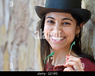 Jeune belle femme de charme Latina porte un chapeau noir, tient des lunettes de soleil et des sourires à la visionneuse devant un fond de mur de pierre. Banque D'Images