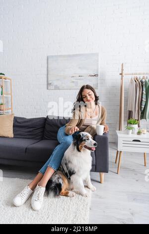 bonne jeune femme assise sur un canapé et câliner le berger australien tout en tenant une tasse de café Banque D'Images