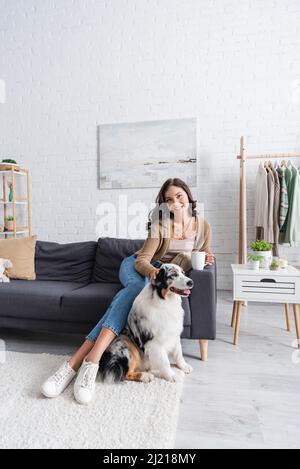 une jeune femme heureuse assise sur un canapé et un chien berger australien en cuddling tout en tenant une tasse de café Banque D'Images