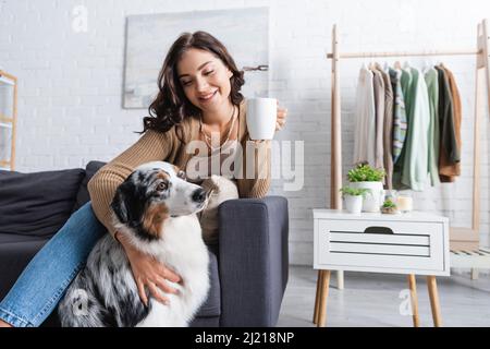 jeune femme joyeuse qui câlin le berger australien et tient une tasse de café Banque D'Images